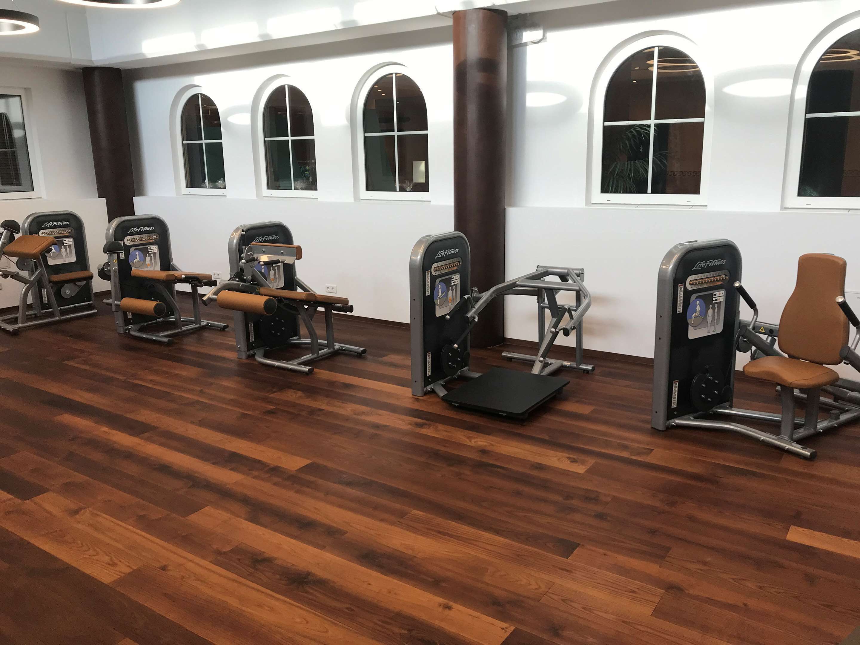 Un gimnasio con varias máquinas de cardio, piso de madera y amplias ventanas arqueadas.