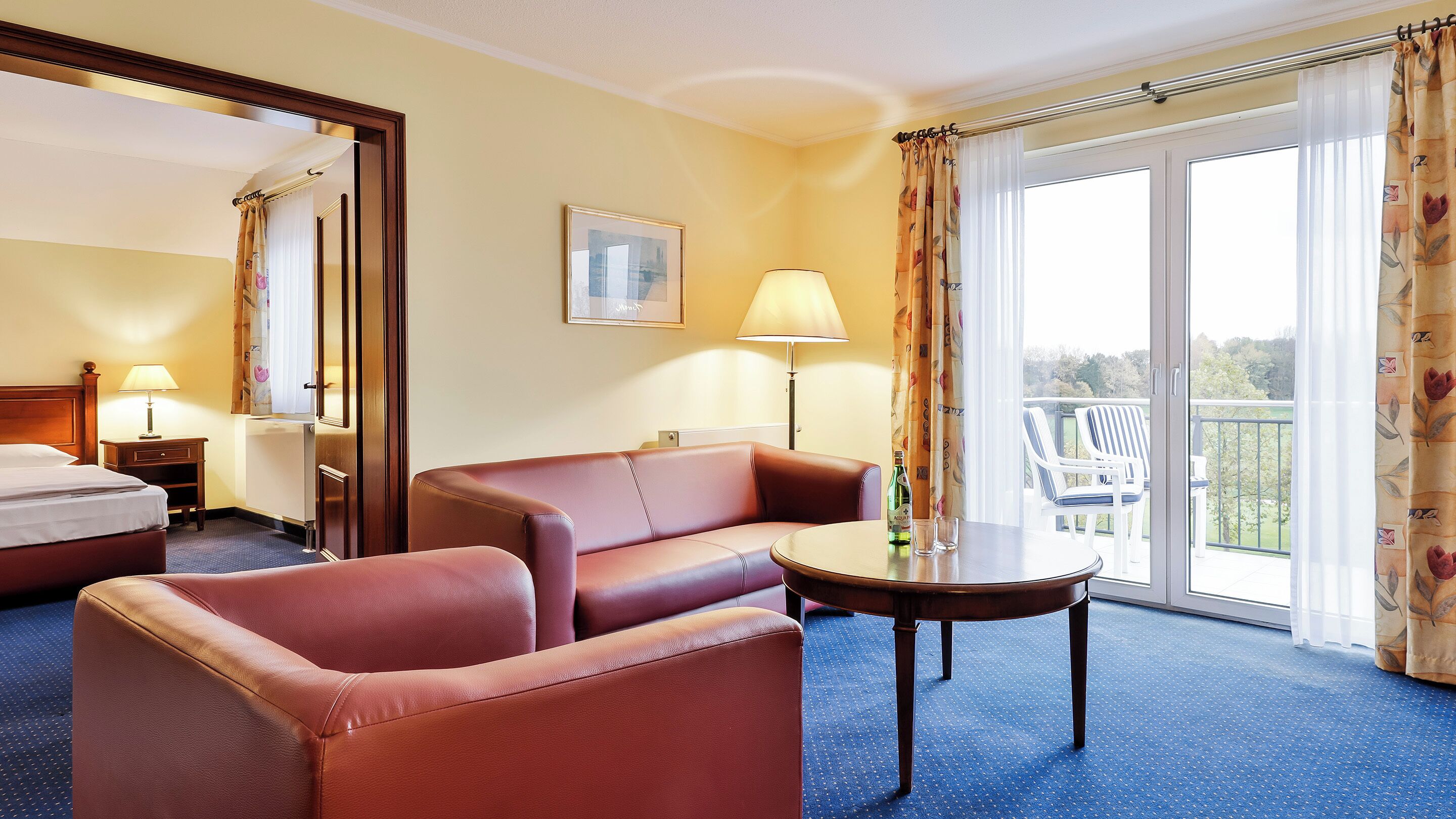 Una habitación de hotel con cama, sofá, mesa, lámpara y balcón con sillas.