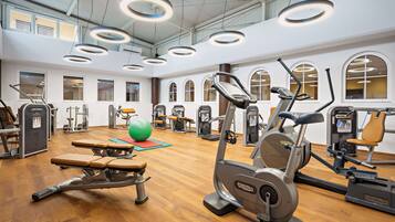 Ein Fitnessraum mit unterschiedlichen Trainingsgeräten, Holzboden und großen Fenstern.