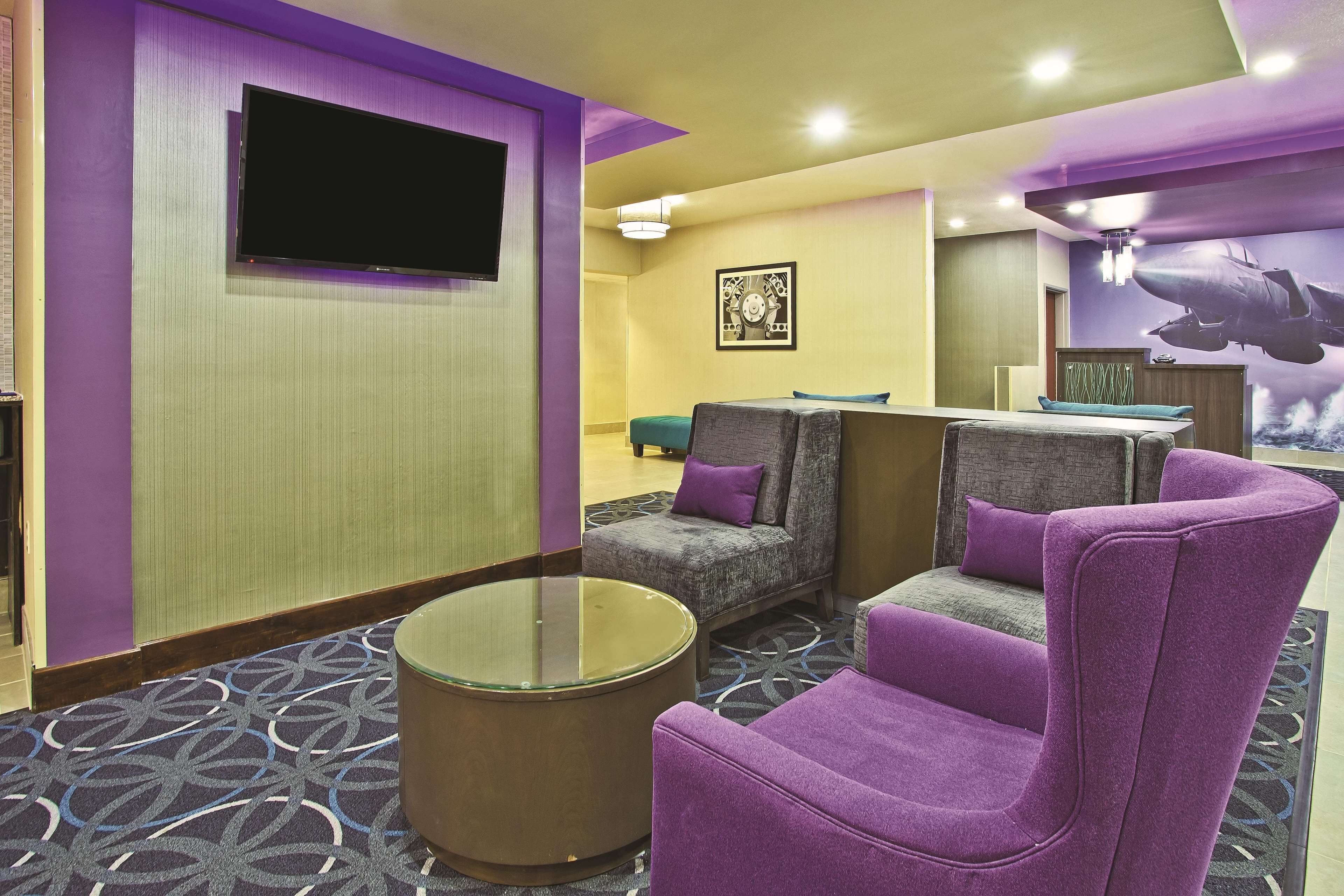 A hotel room with a flat-screen TV mounted on the wall, a round glass coffee table, and two purple armchairs.
