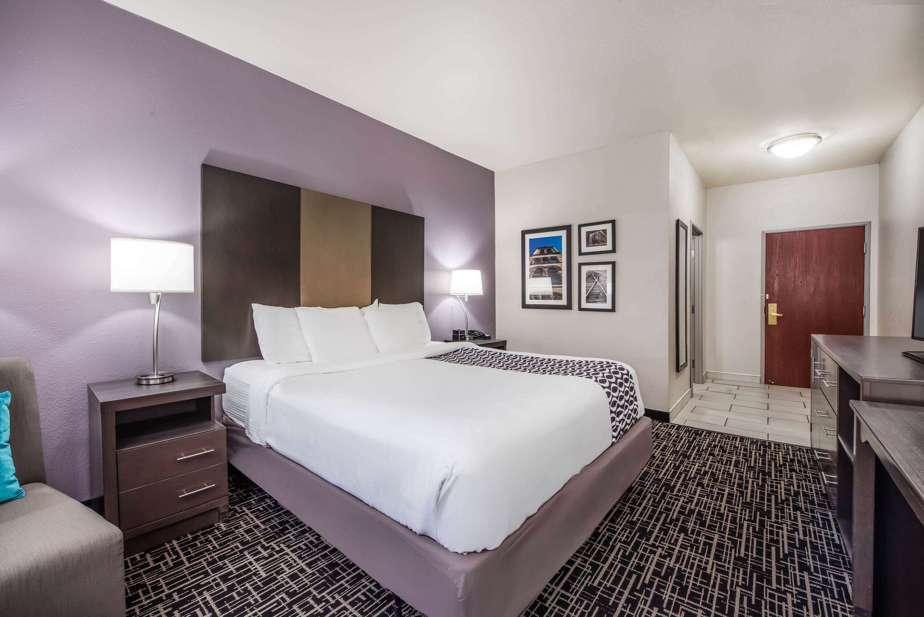 A hotel room with a large bed, a nightstand with a lamp, a desk with drawers, and a door.