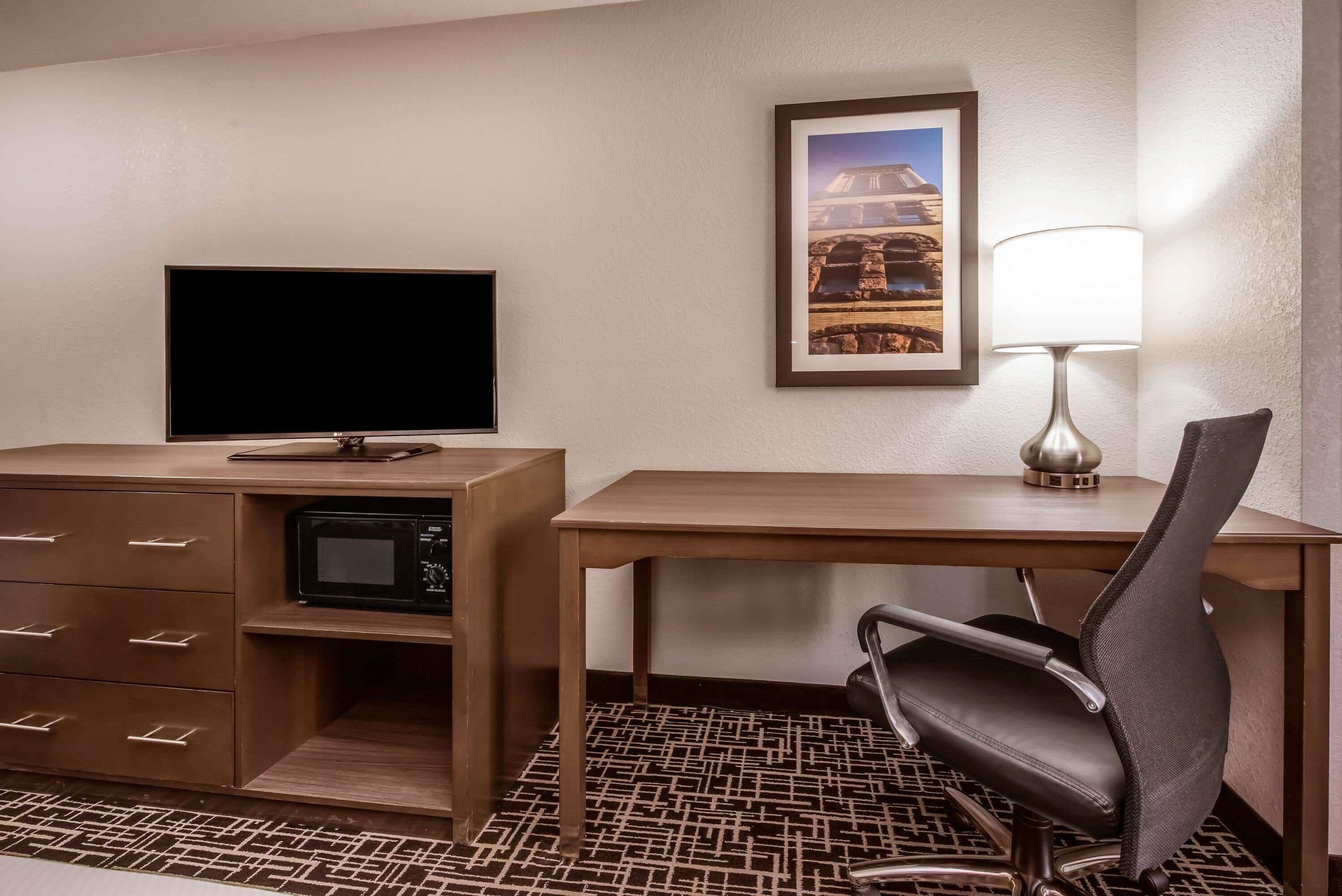 A hotel room with a desk, a television, a microwave, a chair, a lamp, and a framed picture on the wall.