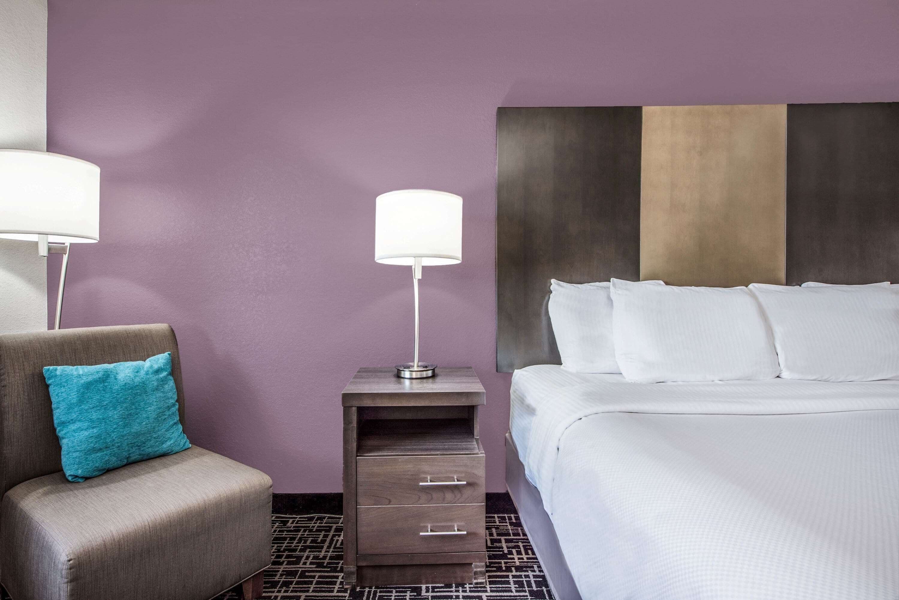 A hotel room with a bed, a nightstand with a lamp, a chair with a pillow, and a wall with a textured design.