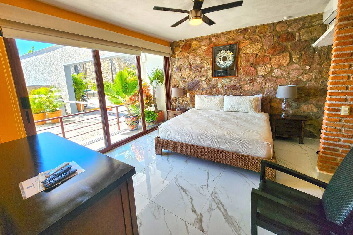 A bedroom with a bed, a ceiling fan, a stone wall, and a view of the outside.