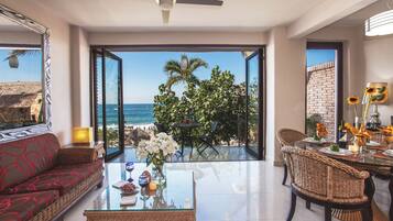 Een ruime woonkamer met een grote glazen deur die uitkomt op een balkon met uitzicht op zee en palmbomen.