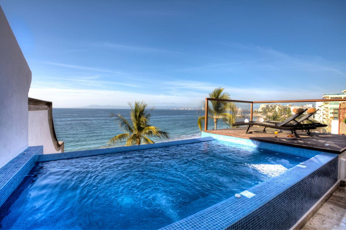 A pool with a view of the ocean, lounge chairs, and palm trees.