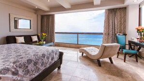 A hotel room with a large bed, a sitting area with a chair and table, a view of the sea, and a desk with a vase of flowers. - Vallarta Shores Beach Hotel (Puerto Vallarta)
