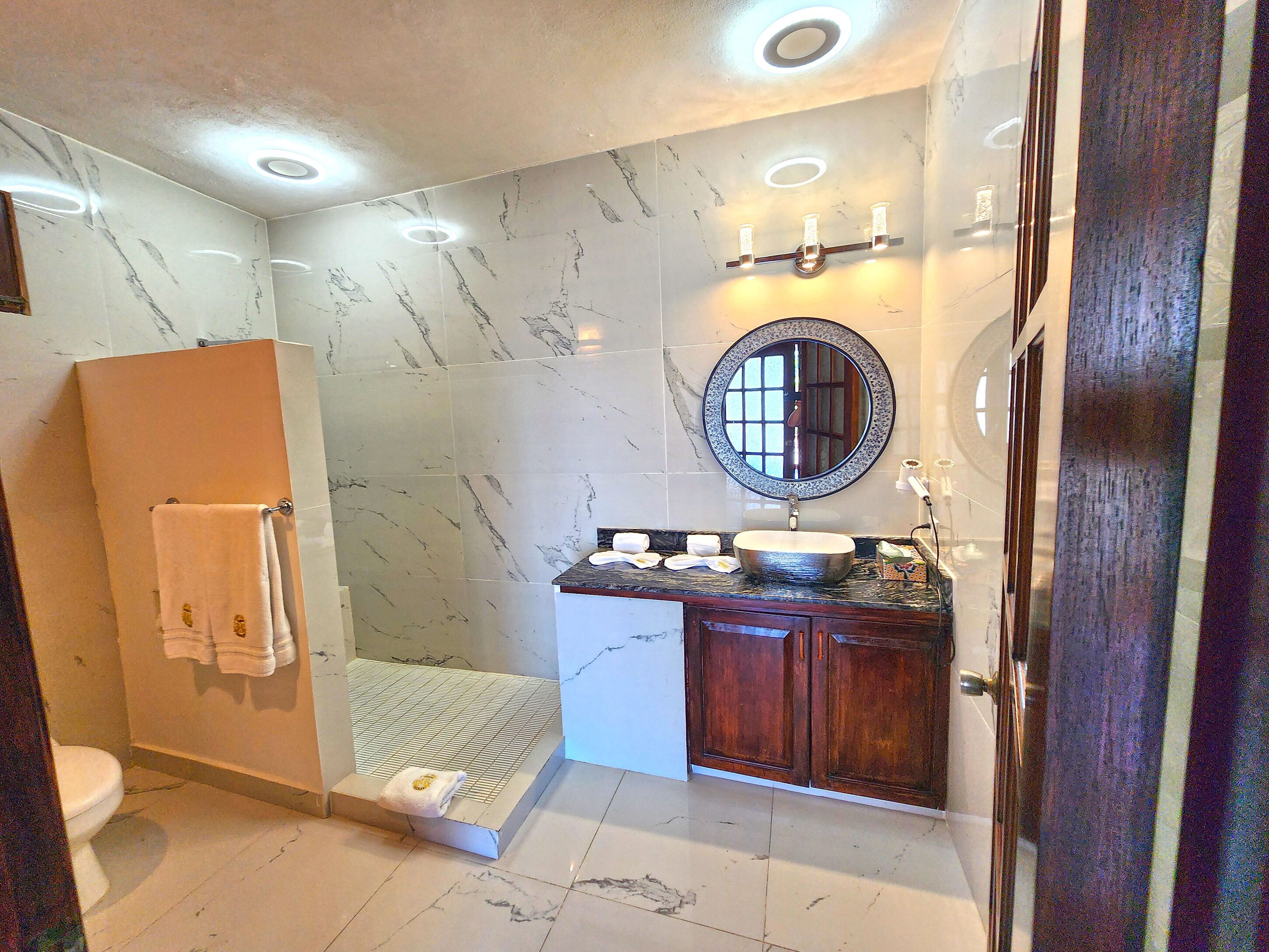 Un baño con piso de mármol, tocador de madera, espejo redondo y cabina de ducha de vidrio.