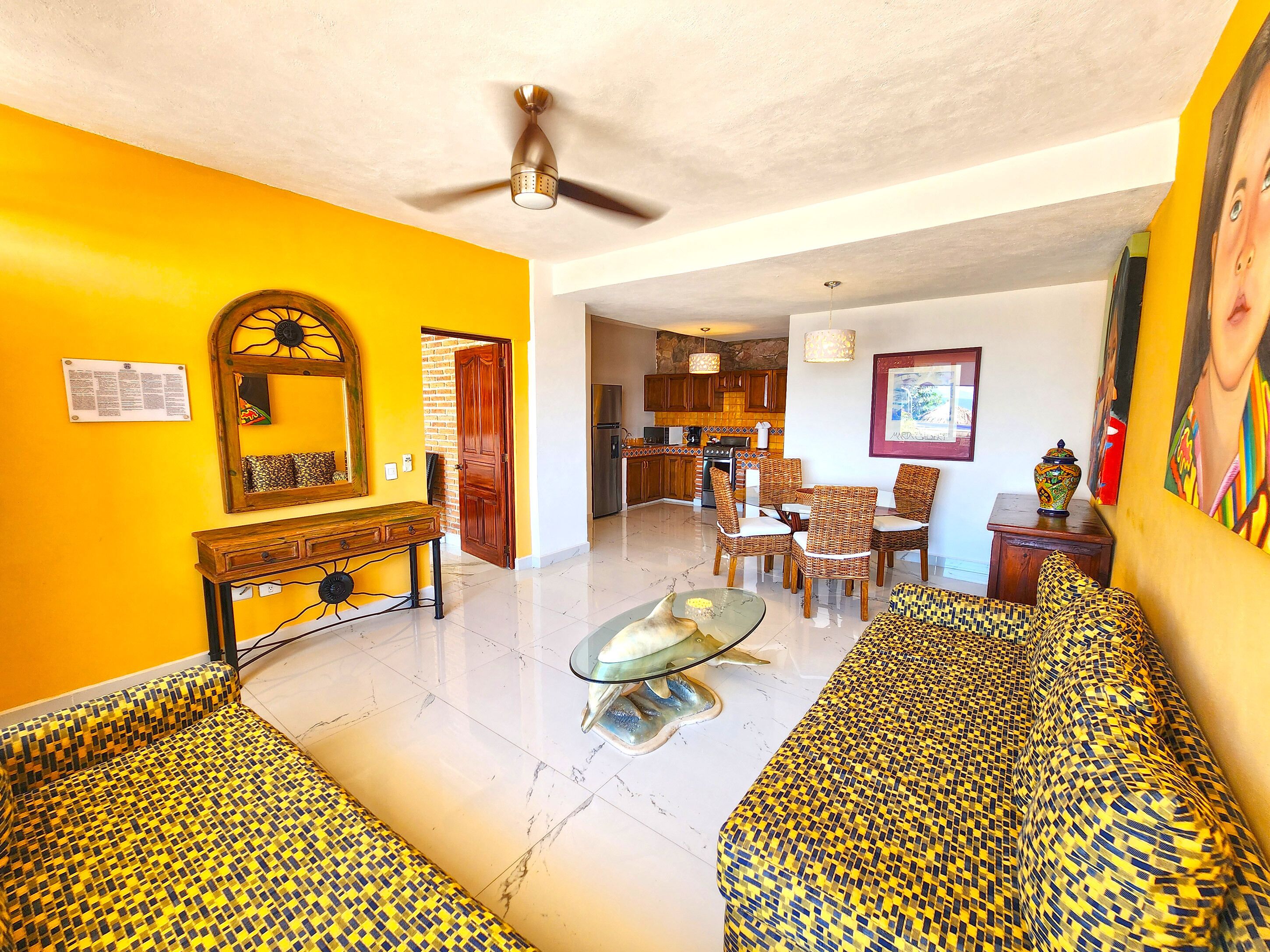 A spacious living area with a yellow wall, a ceiling fan, a dining set, and a kitchen in the background.