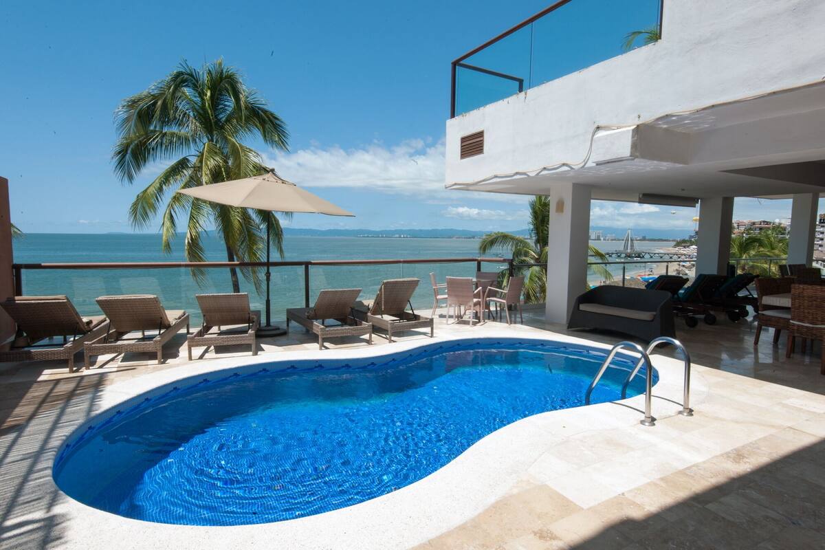 A swimming pool with lounge chairs and umbrellas, overlooking a beach and ocean.