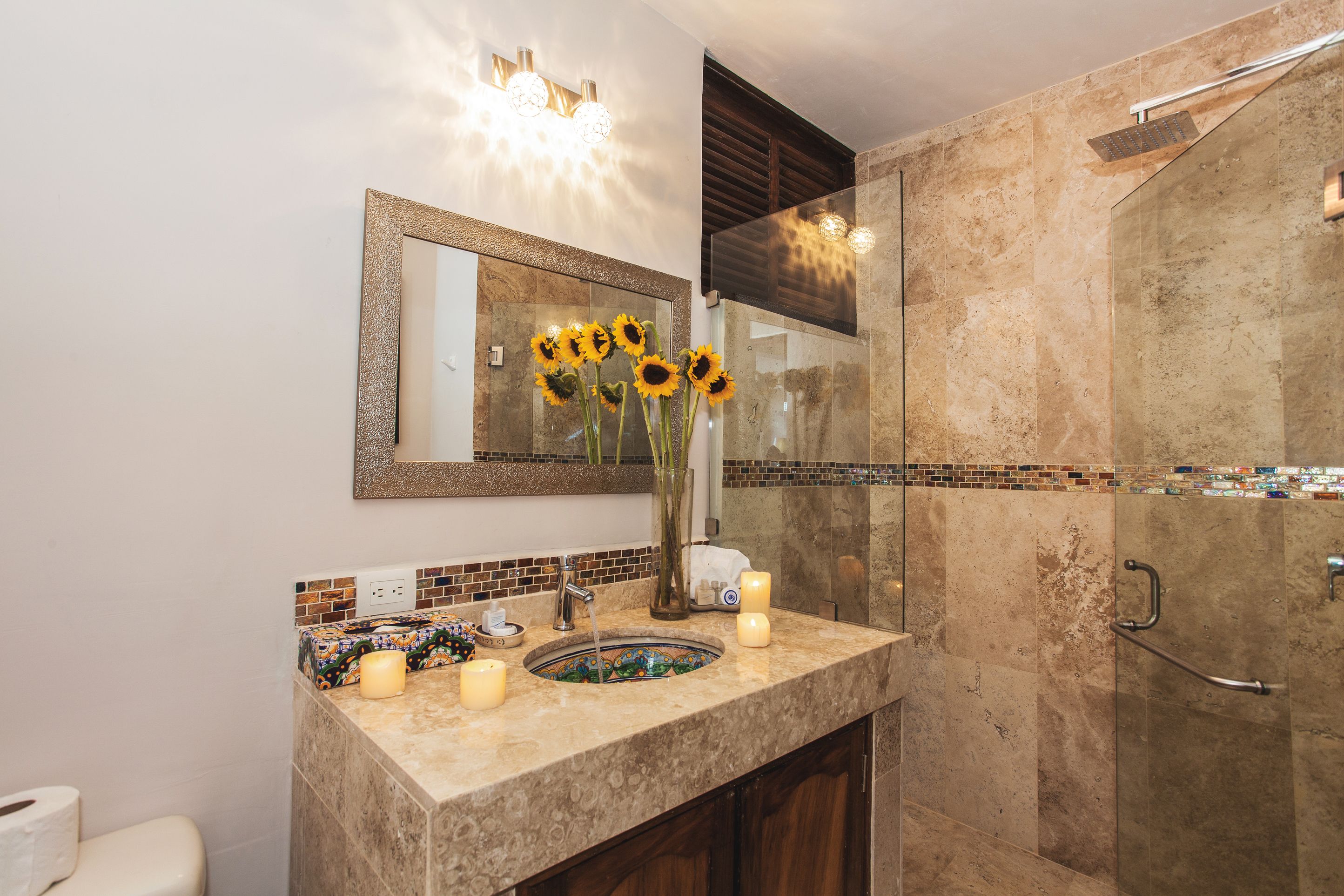 A bathroom with a marble sink, a mirror, a vase with sunflowers, and a glass shower enclosure.