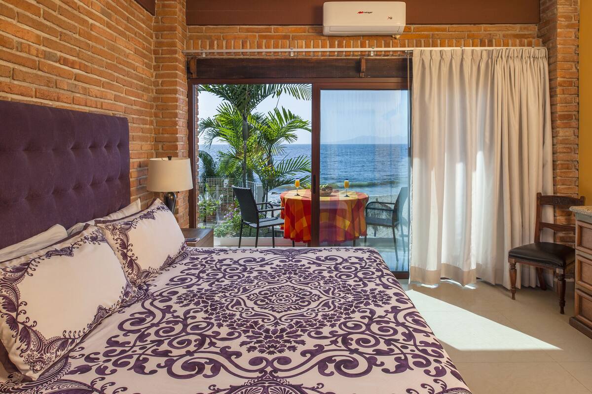 A hotel room with a large bed, a view of the ocean, a balcony with a table and chairs, and a brick wall.