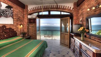 A bedroom with a view of the ocean, a bed with green bedding, a dresser, and a TV.