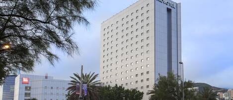 Un hotel de gran altura con un llamativo letrero de 'Novotel', rodeado de palmeras y otros edificios.
