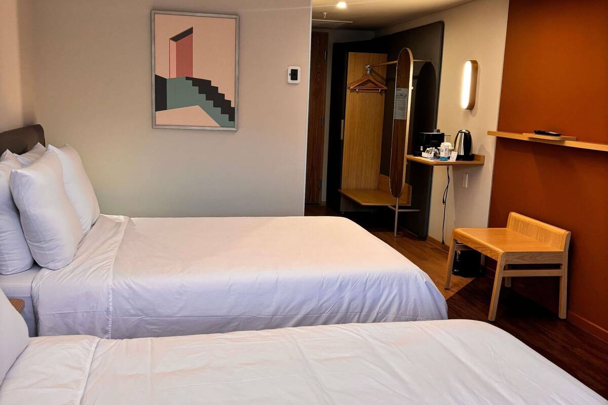 A hotel room with two beds, a wooden chair, a desk with a coffee maker, a mirror, and a door leading to a bathroom.