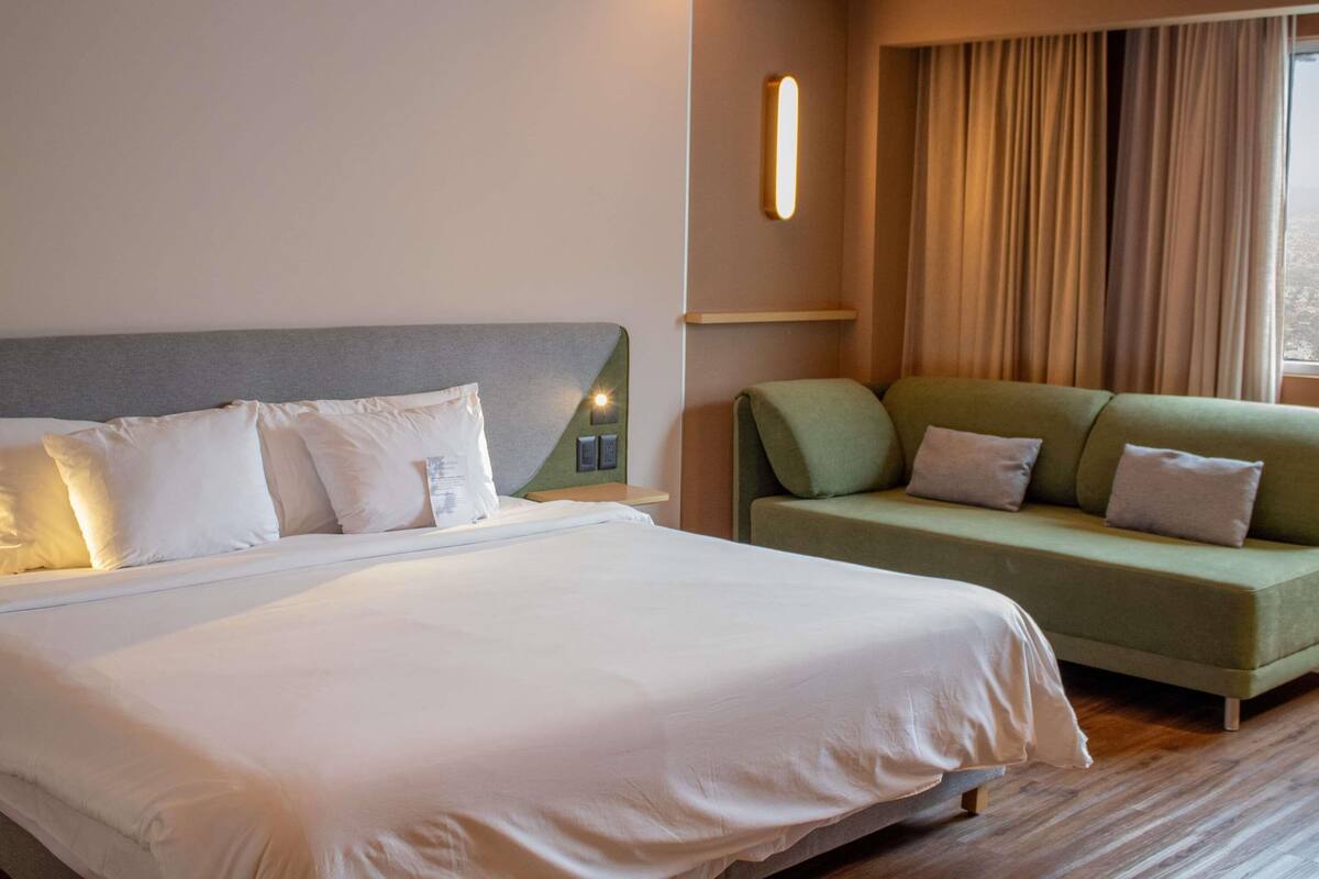 A hotel room with a bed, a green sofa, a wall-mounted light, and a window with curtains.