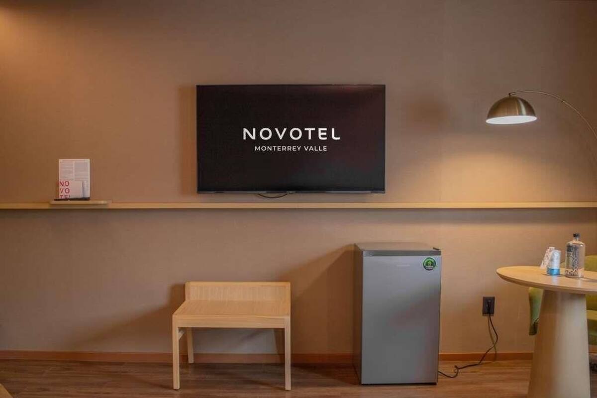A hotel room with a TV, a small refrigerator, a bench, and a table with a lamp.
