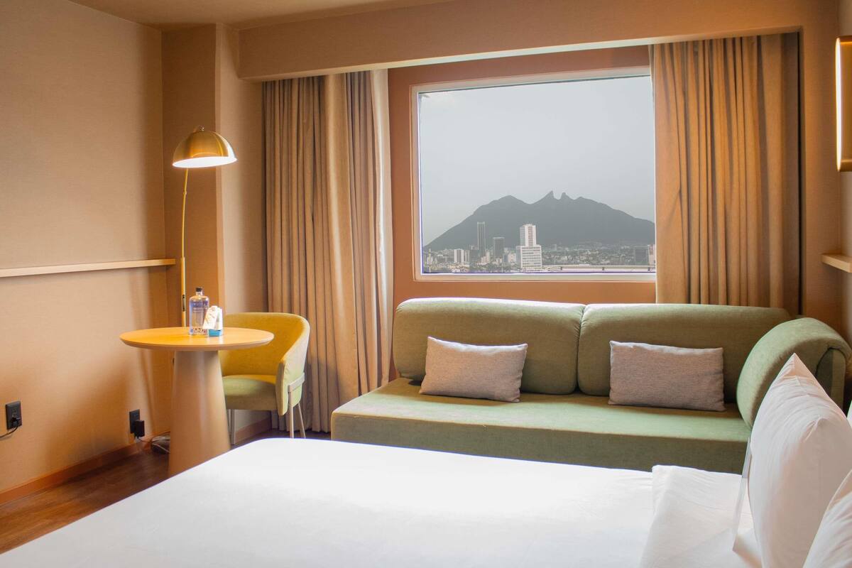 A hotel room with a bed, a sofa, a small table, and a view of a cityscape and mountain.
