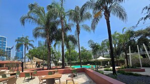 An outdoor seating area with tables and chairs, surrounded by palm trees and a swimming pool. - Novotel Monterrey Valle (San Pedro Garza García)