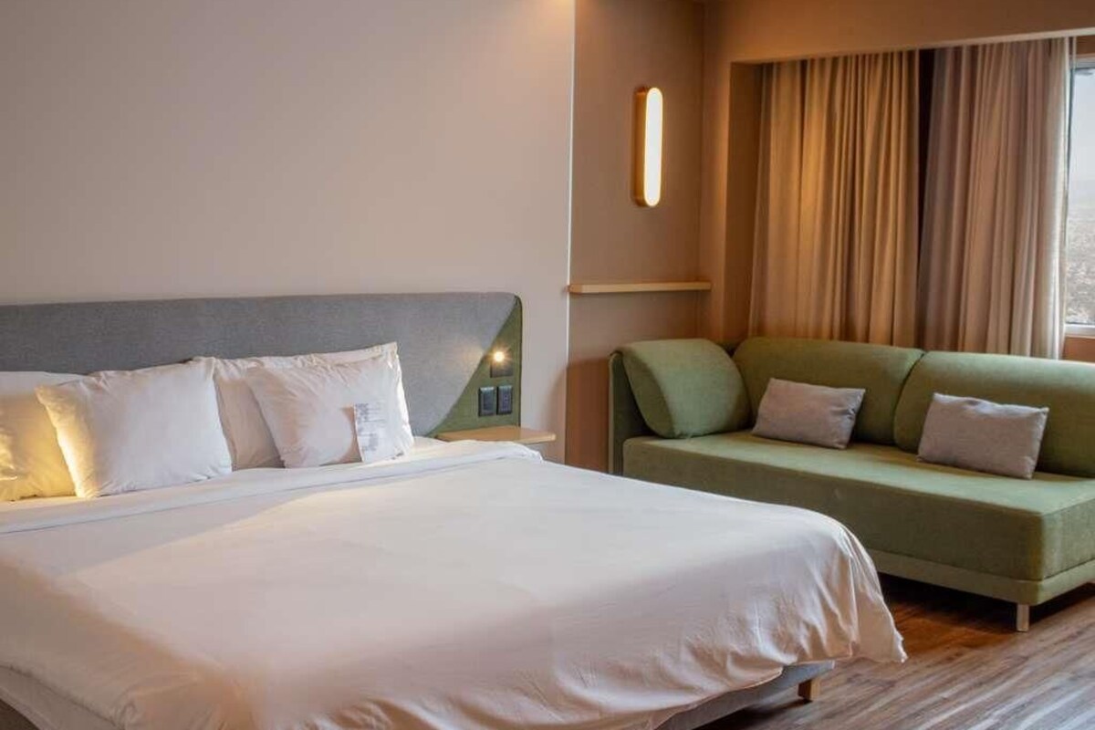 A hotel room with a large bed, a green sofa, and a window with curtains.