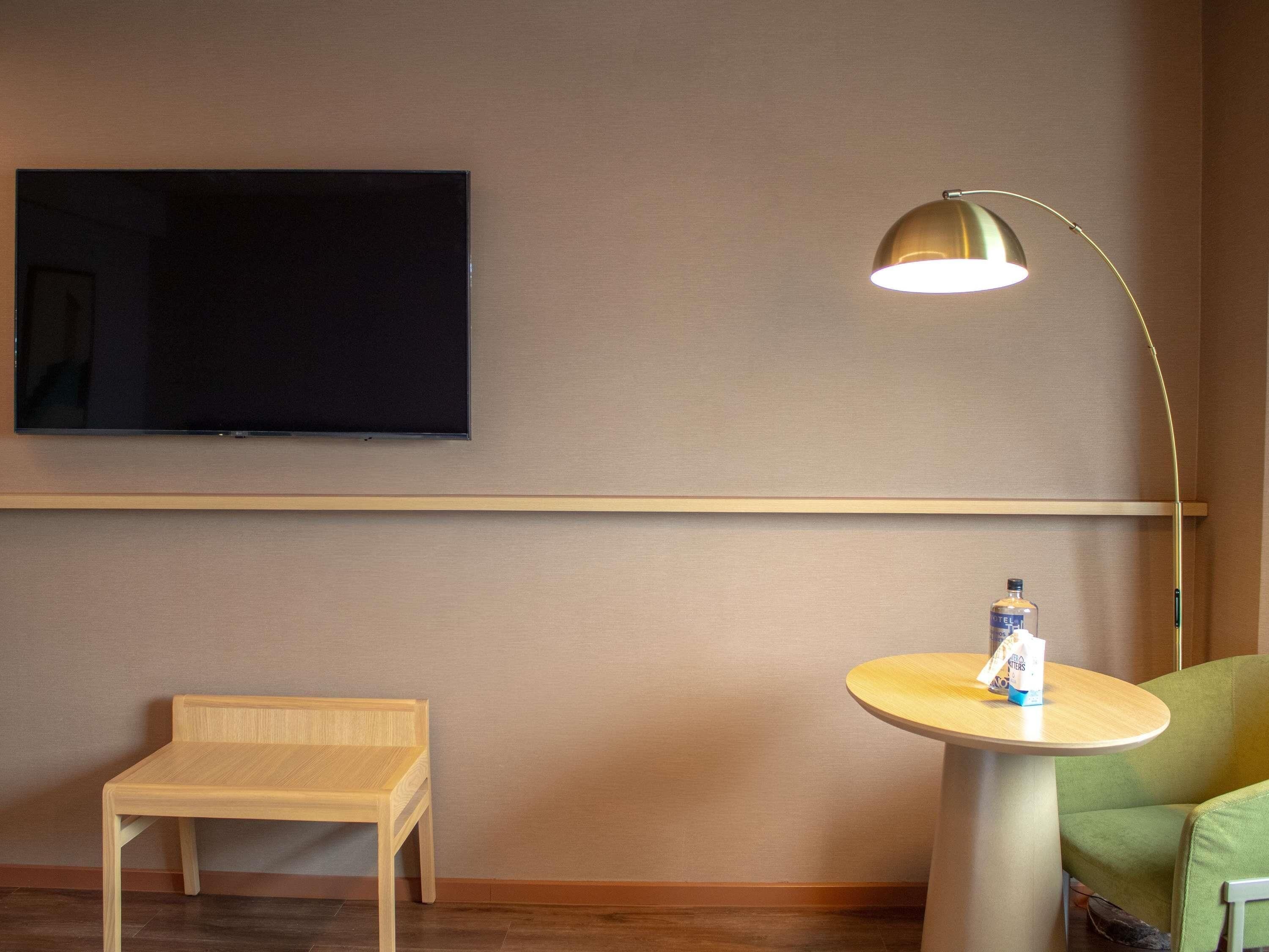 Une pièce avec un téléviseur fixé au mur, une petite table en bois, une table ronde avec une bouteille et une chaise verte.