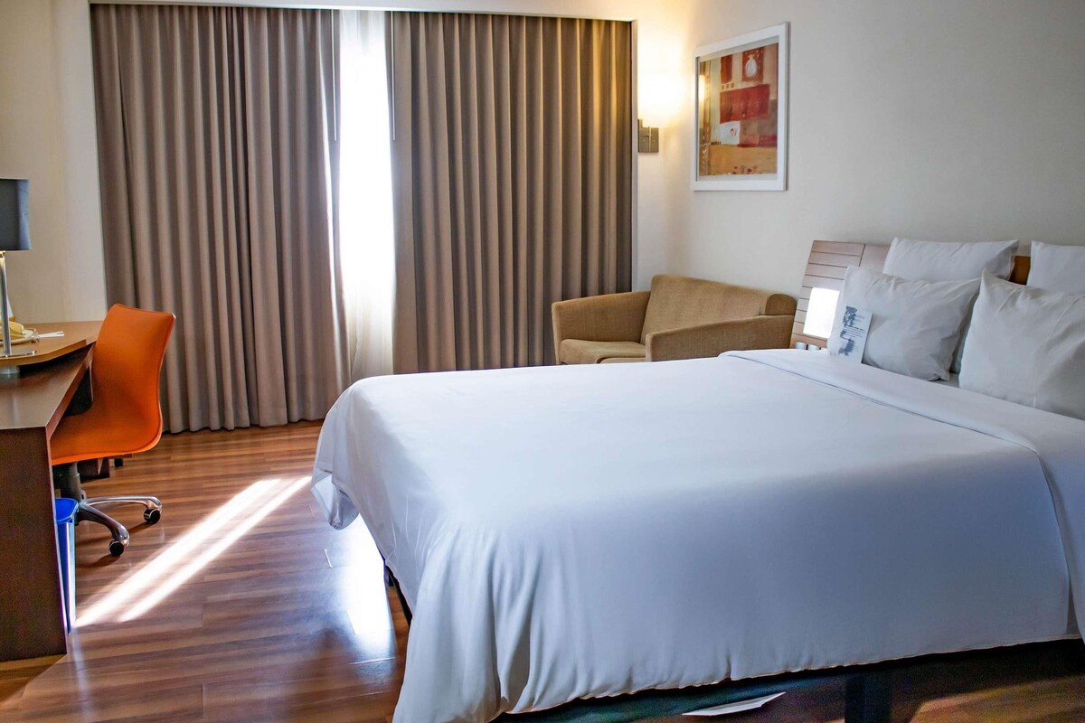 A hotel room with a large bed, a desk with a chair, a sofa, and a painting on the wall.