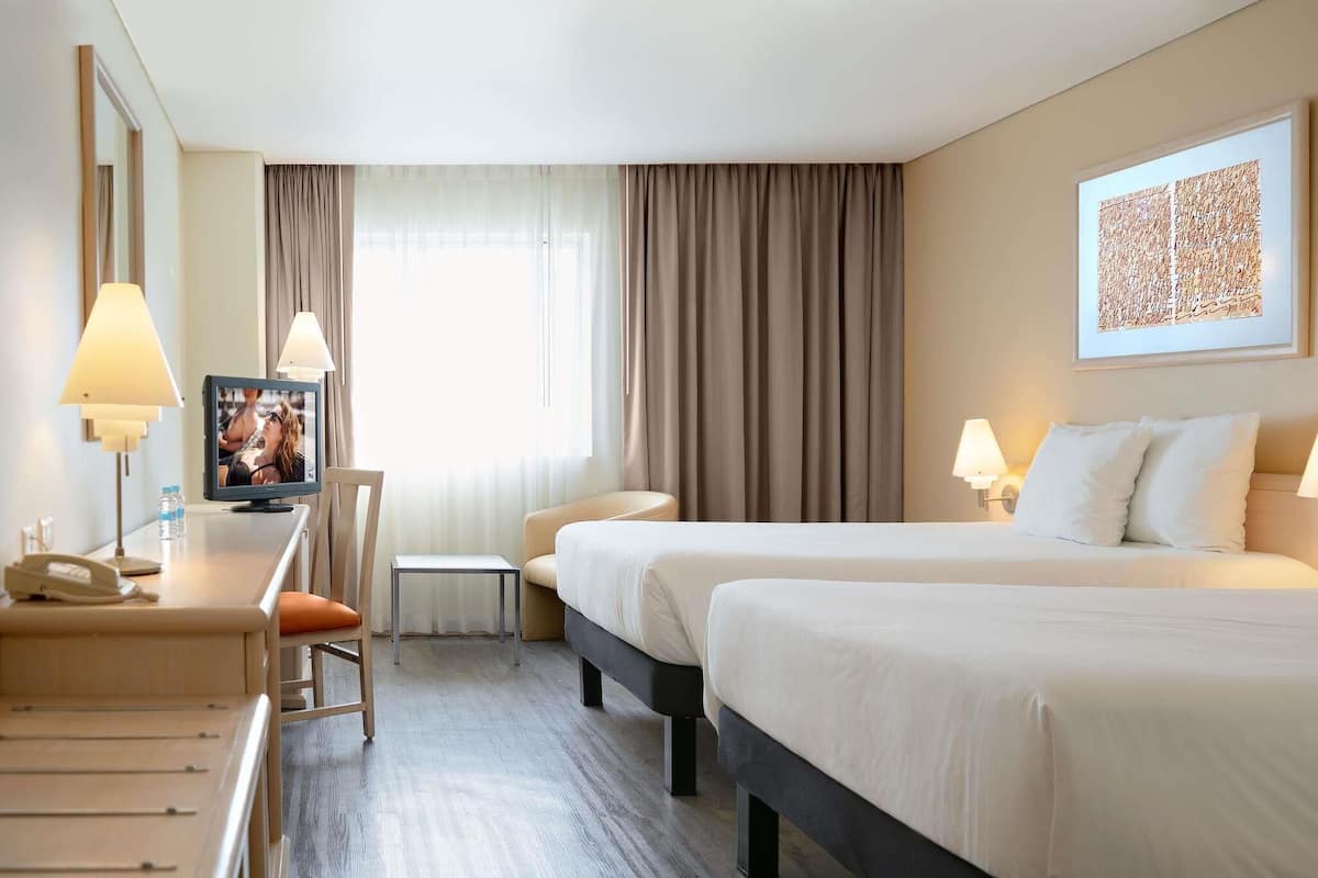 A hotel room with two beds, a desk with a lamp, a chair, a small table, and a window with curtains.