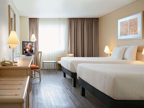 Une chambre d’hôtel avec deux lits, un bureau avec une lampe, une chaise, une petite table et une fenêtre avec des rideaux.