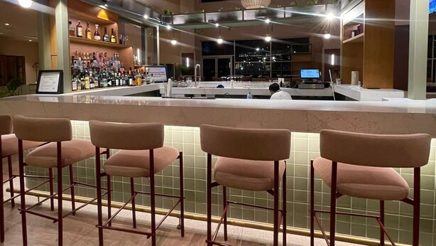 A bar area with a long counter, multiple bar stools, hanging plants, and pendant lighting. - Novotel Monterrey Valle (San Pedro Garza García)