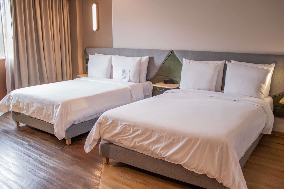 Une chambre d’hôtel avec deux lits, un plancher en bois, une grande fenêtre avec des rideaux et un luminaire fixé au mur.
