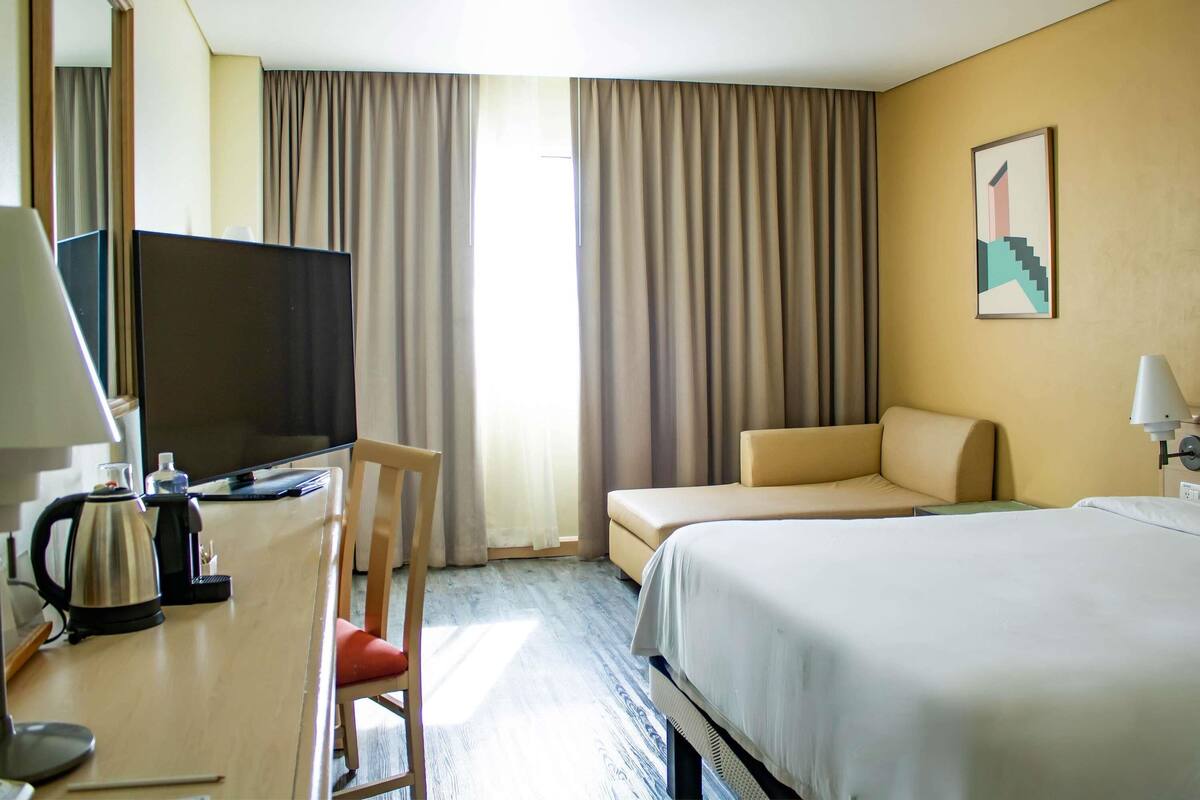 A hotel room with a bed, a desk with a television, a chair, a sofa, and a painting on the wall.
