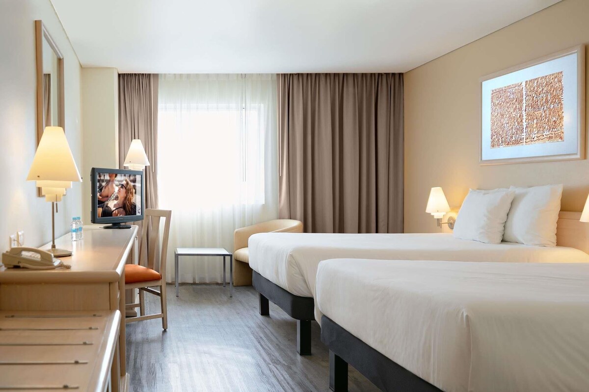 Une chambre d’hôtel avec deux lits, un bureau avec une lampe, une chaise, une petite table et une fenêtre avec des rideaux.