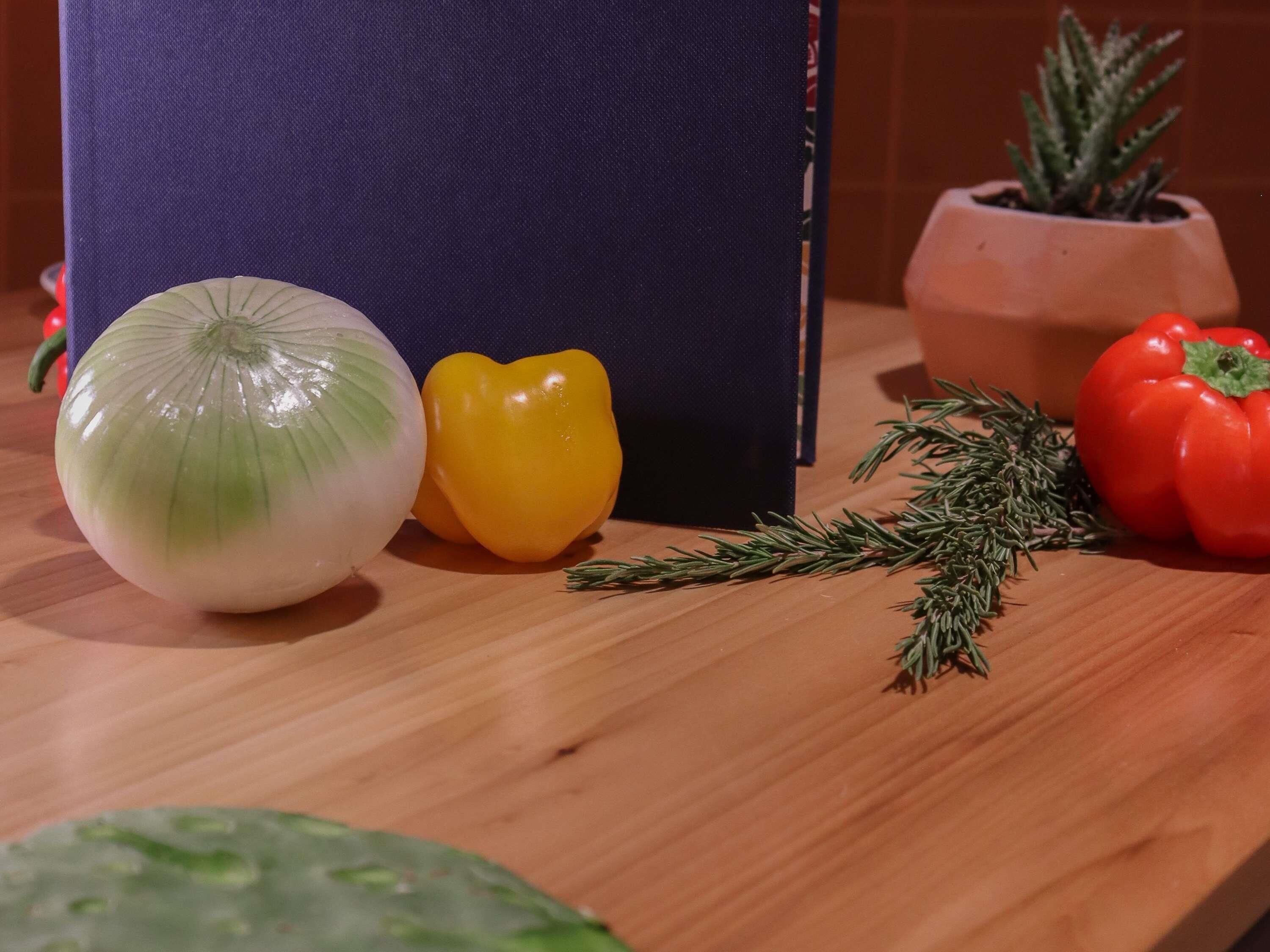 A kitchen counter with a white onion, a yellow bell pepper, a red bell pepper, a sprig of rosemary, and a potted plant.