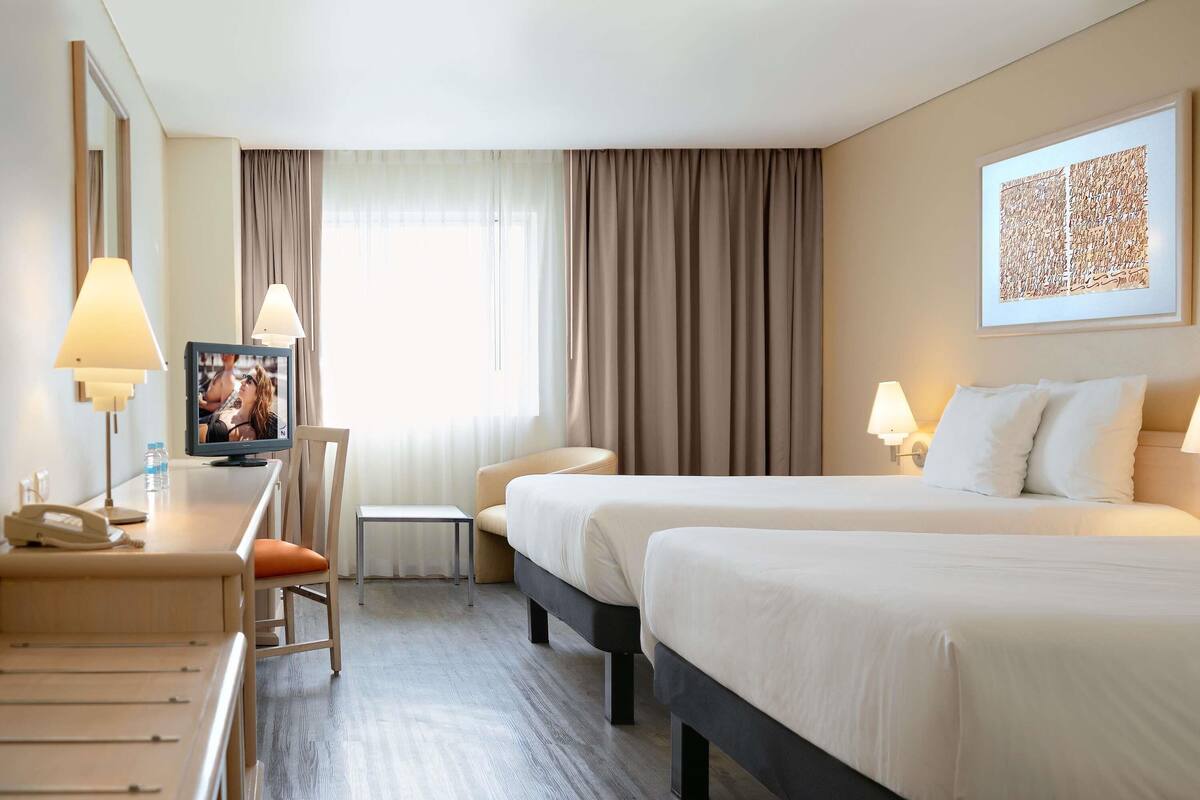 A hotel room with two beds, a desk with a phone, a chair, a TV, and a window with curtains.