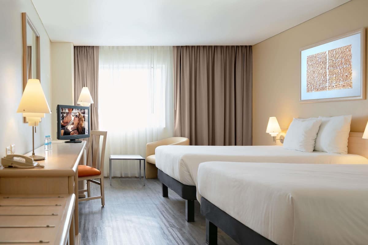 A hotel room with two beds, a desk with a phone, a chair, a TV, and a window with curtains.