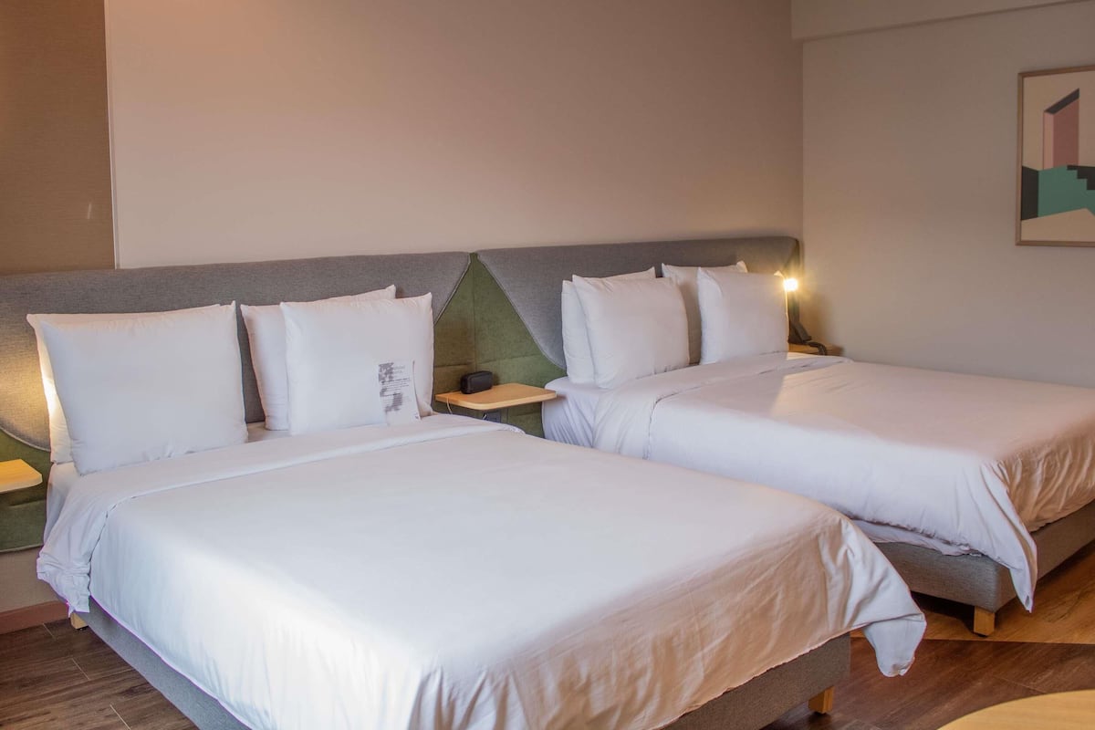 Quarto de hotel com duas camas, uma mesa de cabeceira com abajur e uma obra de arte na parede.