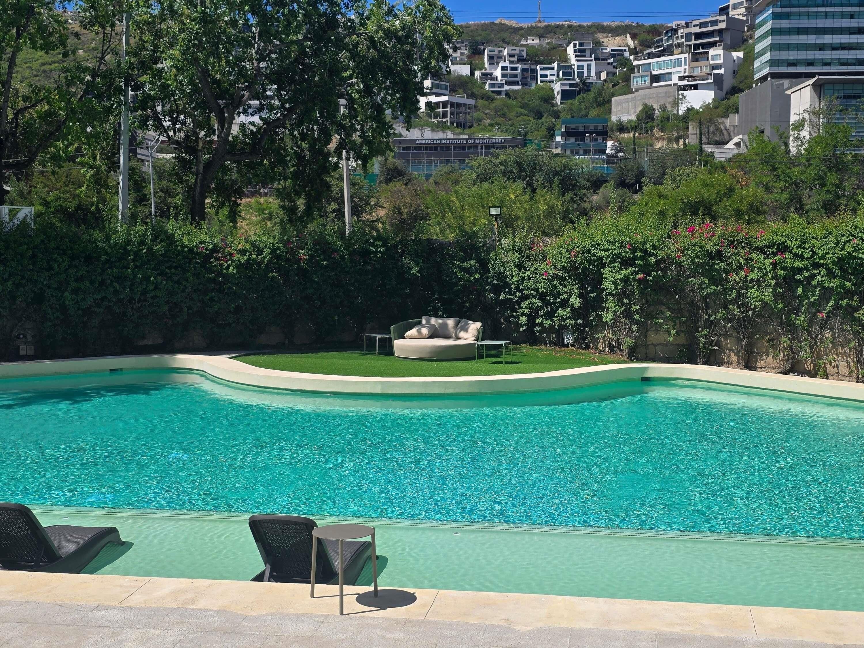 A swimming pool with lounge chairs and a small table, surrounded by greenery and buildings in the background.