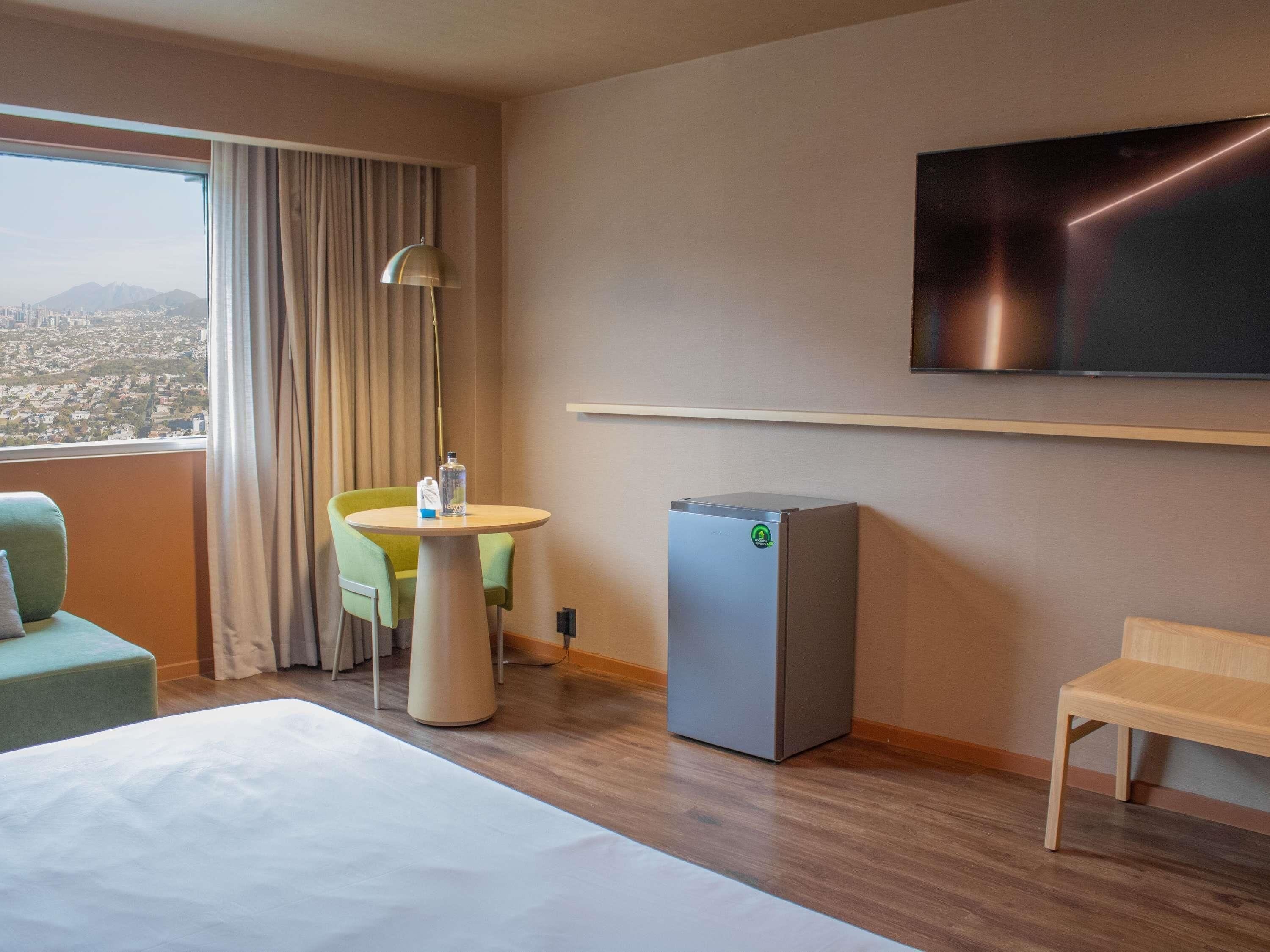 A hotel room with a bed, a small table with a chair, a refrigerator, a TV mounted on the wall, and a window with curtains.