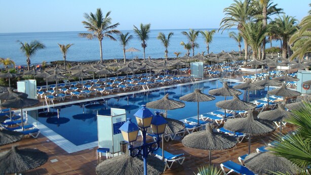 Ein Poolbereich mit Liegestühlen und Sonnenschirmen, Palmen und Blick aufs Meer.