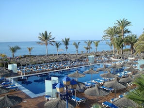 A pool area with lounge chairs and umbrellas, palm trees, and a view of the sea. - VIK Hotel San Antonio (Tías)