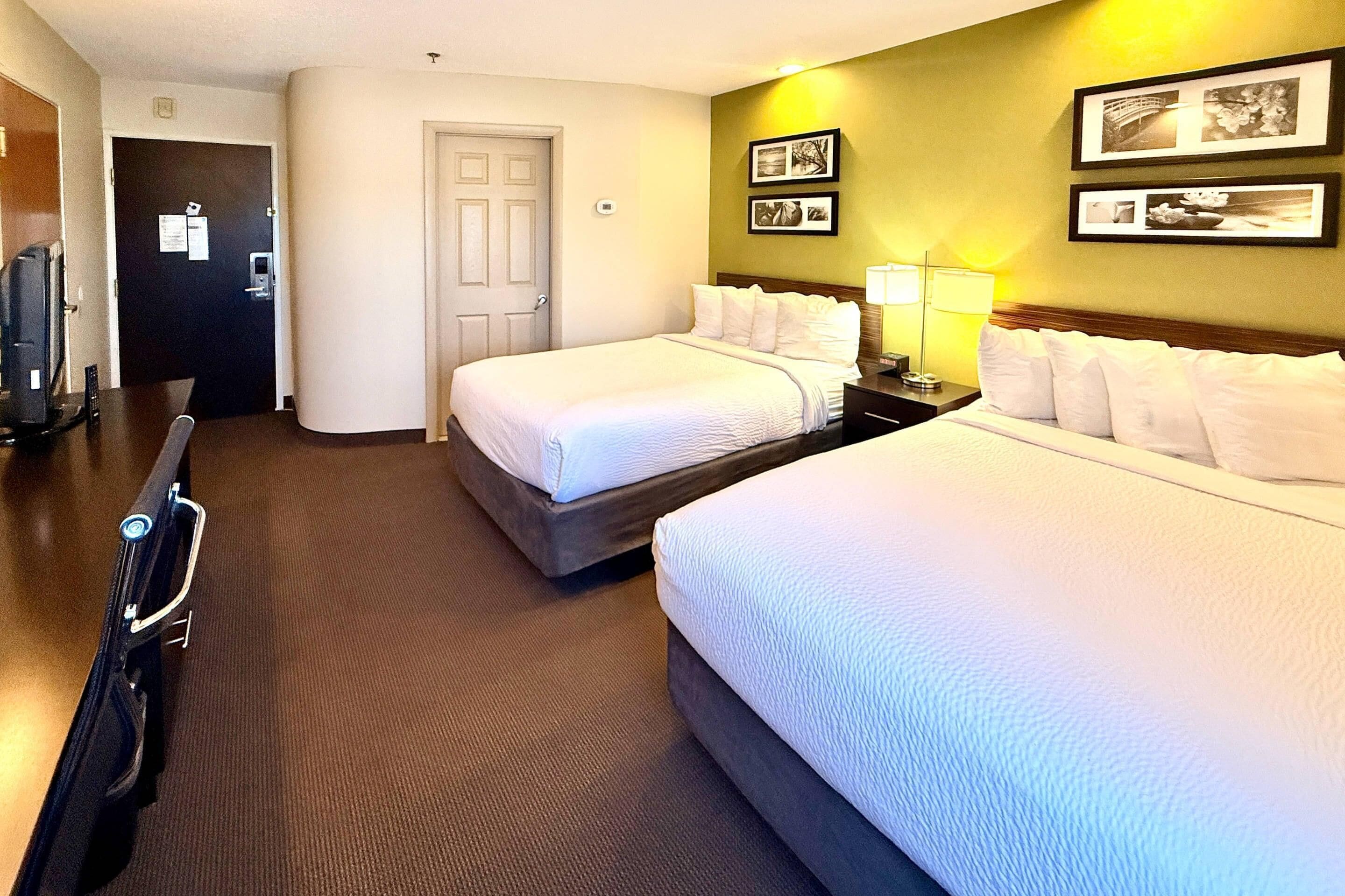 Habitación de hotel con dos camas, un escritorio, una silla, un televisor y cuadros en la pared.