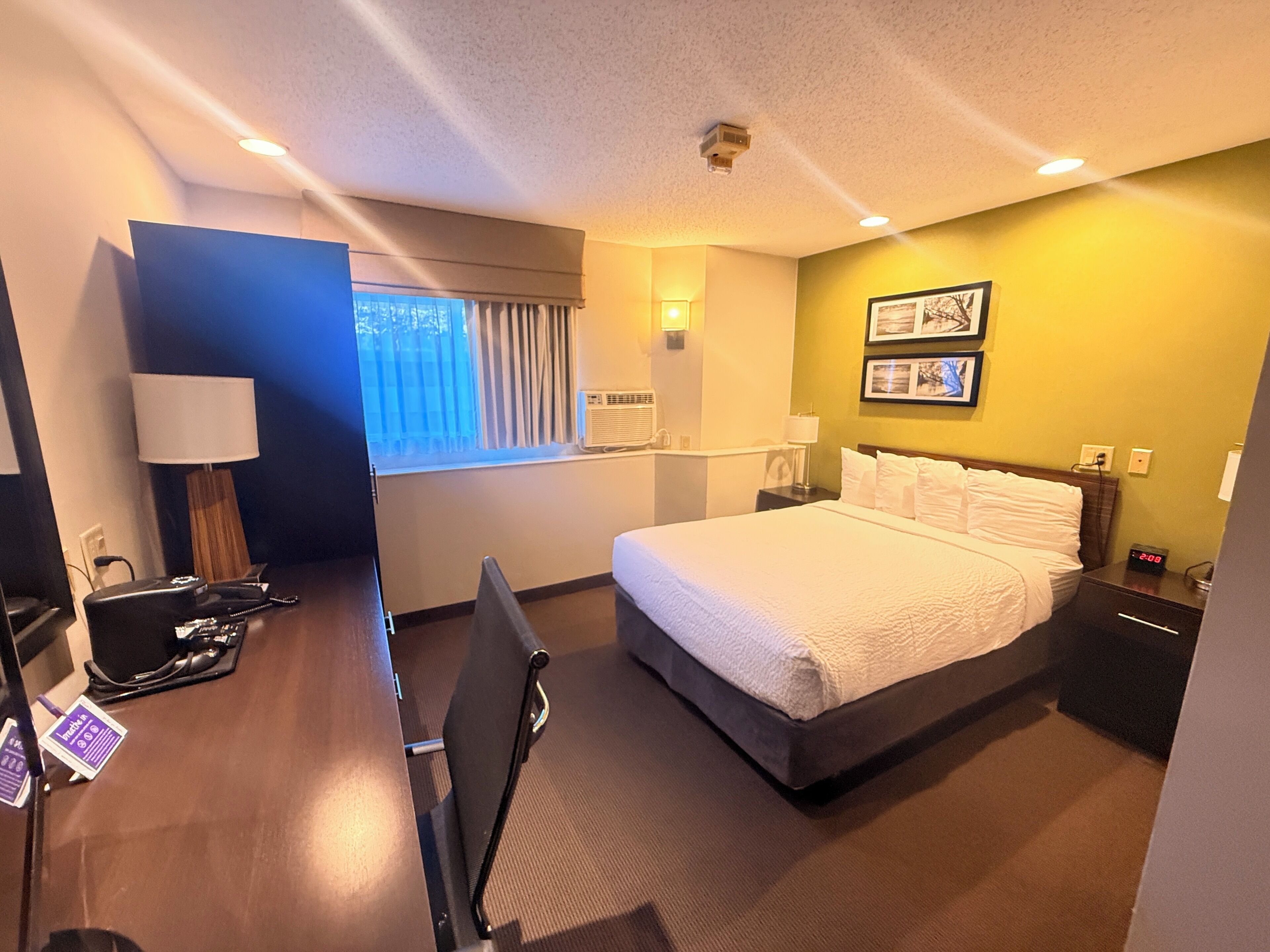 A hotel room with a bed, desk, chair, lamp, bedside table, and wall-mounted air conditioner.