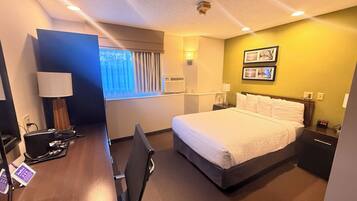 A hotel room with a bed, desk, chair, lamp, bedside table, and wall-mounted air conditioner.