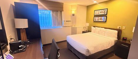 A hotel room with a bed, desk, chair, lamp, bedside table, and wall-mounted air conditioner.