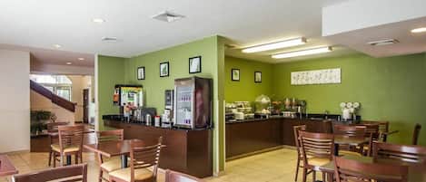 A dining area with wooden tables and chairs, a counter with a refrigerator and coffee machines, and green walls with framed pictures.