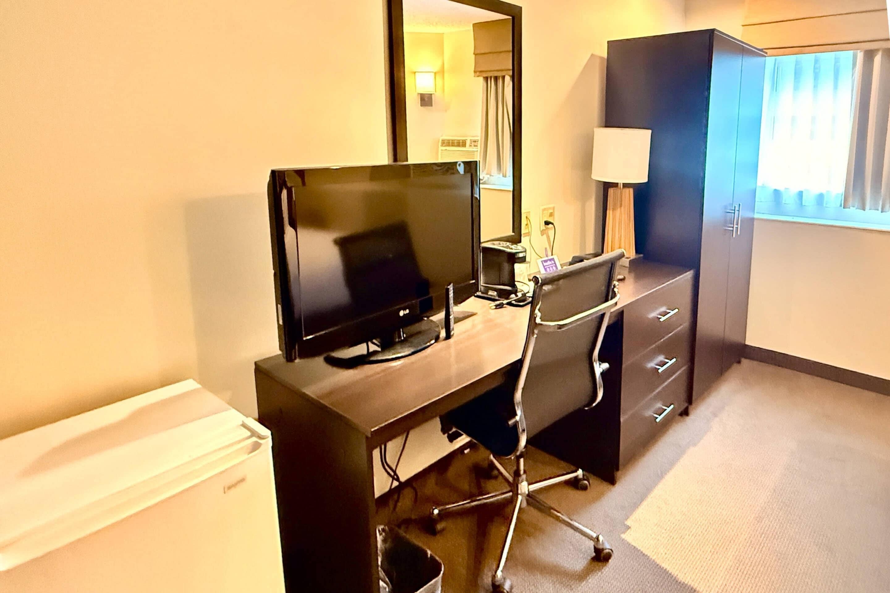 Quarto de hotel com escrivaninha, cadeira, televisão, guarda-roupa e janela com cortinas.