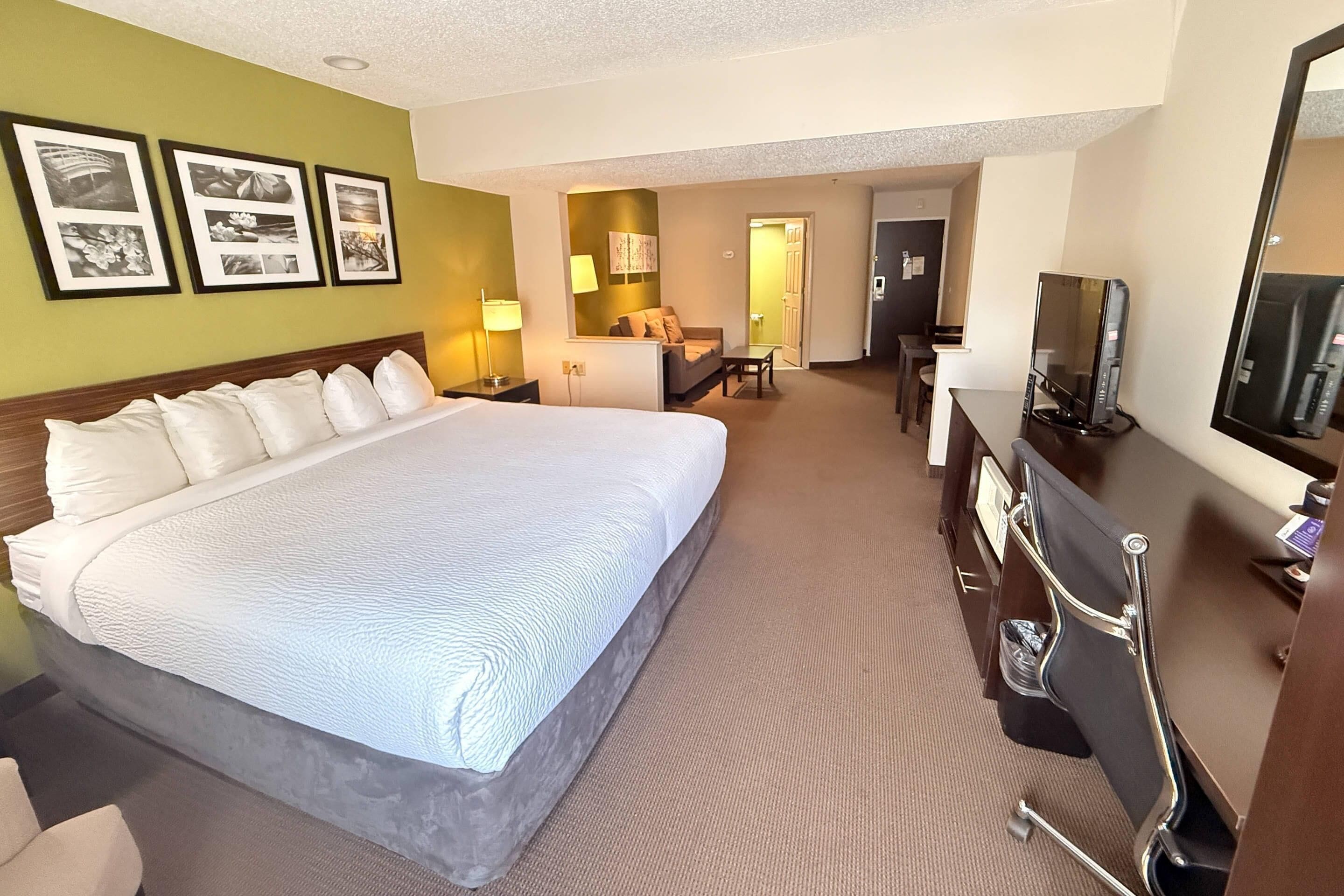 A hotel room with a large bed, a desk with a computer, a TV, and a bathroom visible through an open door.