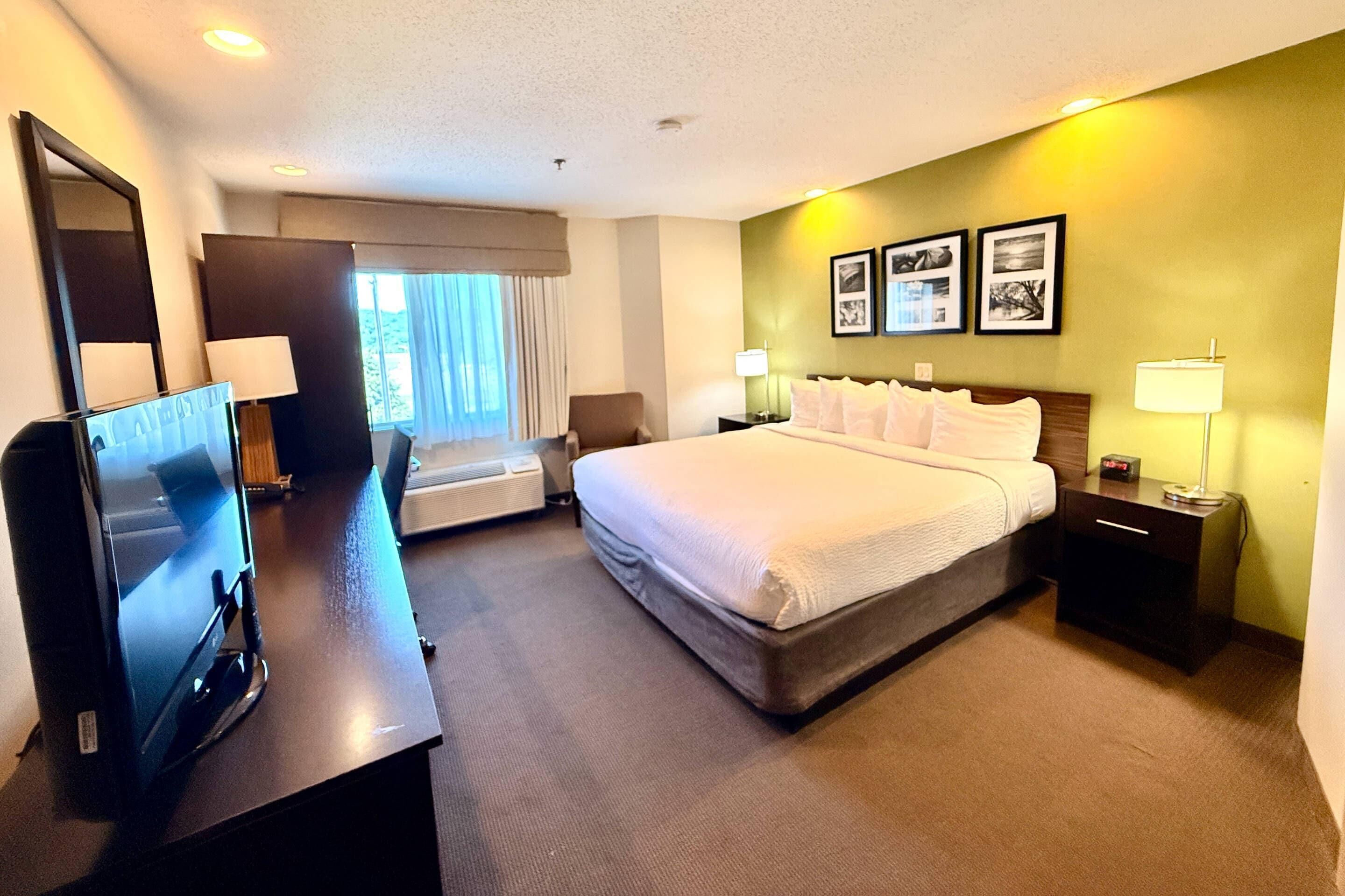A hotel room with a bed, bedside tables, a desk with a computer, a window with curtains, and wall-mounted artwork.