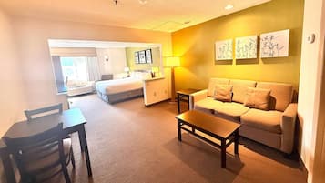 Ein Hotelzimmer mit einem Bett, einem Sofa, einem Tisch und einem Schreibtisch. Zwei Lampen und ein Fenster mit Vorhängen sind vorhanden.