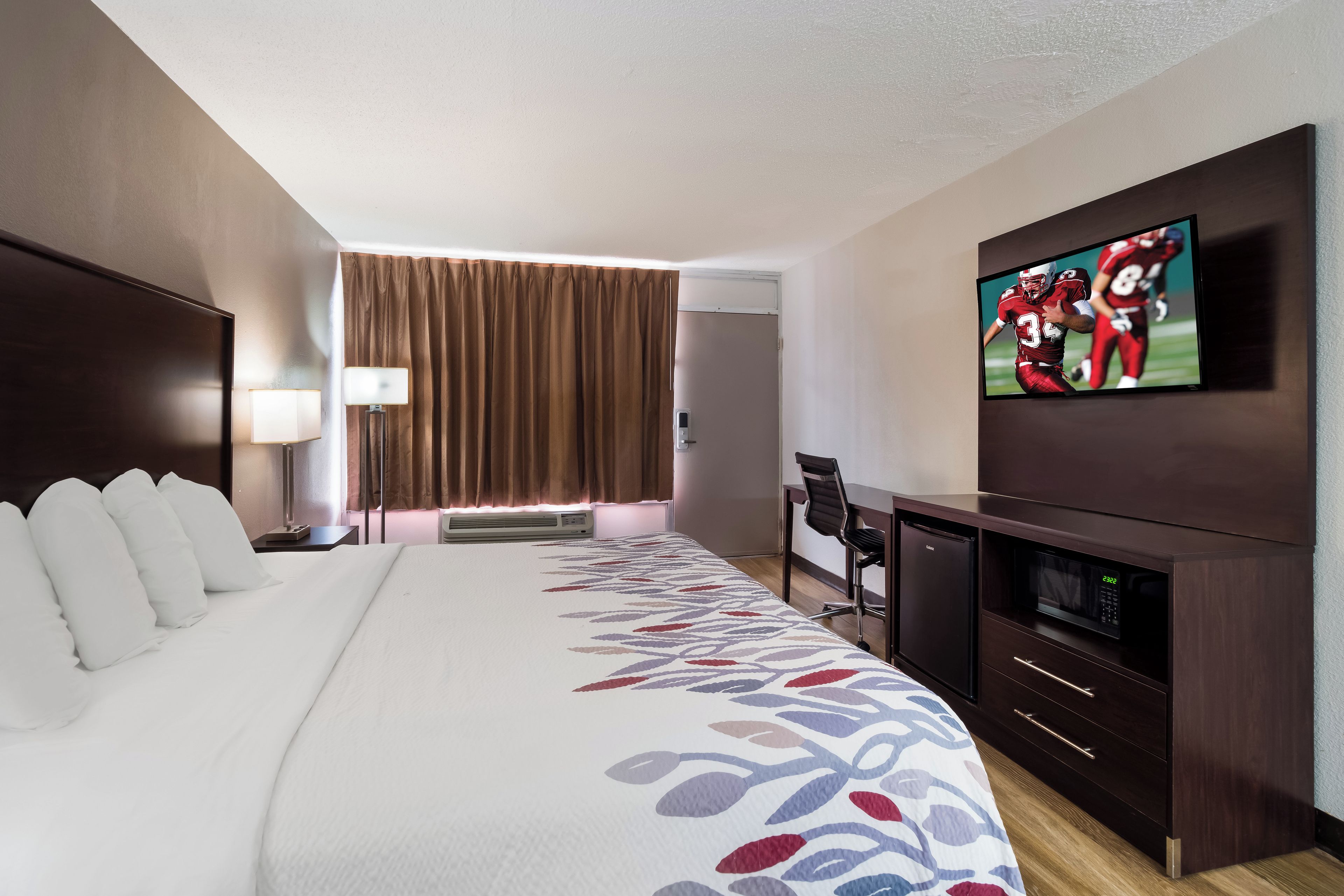 A hotel room with a large bed, a desk with a chair, a television, and an air conditioning unit.